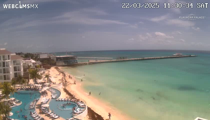 A beach resort with pools overlooks a calm, turquoise ocean and a long pier with a boat under partly cloudy skies.