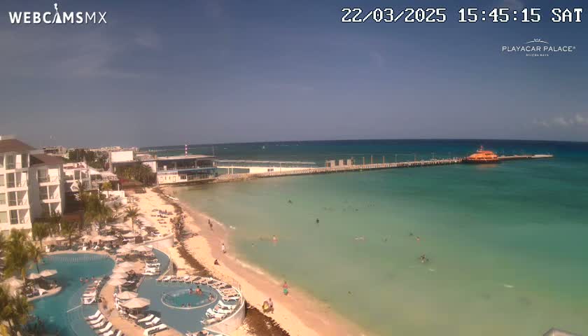 A beach resort with a swimming pool area overlooks a calm, clear turquoise ocean with a pier and boat, under a mostly sunny sky.