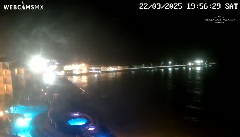 A nighttime view of a resort pool area overlooking a calm, dark ocean with a brightly lit pier in the distance under cloudy skies.