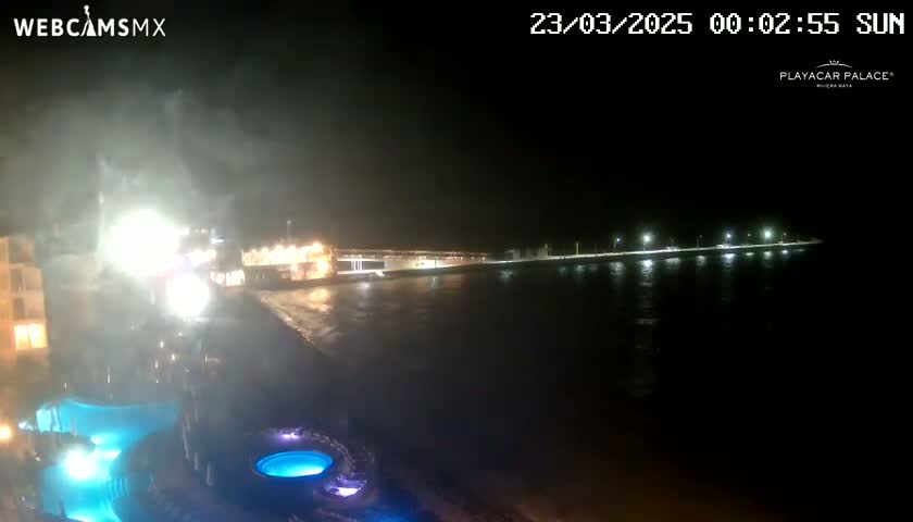 A nighttime coastal scene shows a building with illuminated pools in the foreground and a dimly lit waterfront and shoreline in the background, under hazy or foggy conditions.