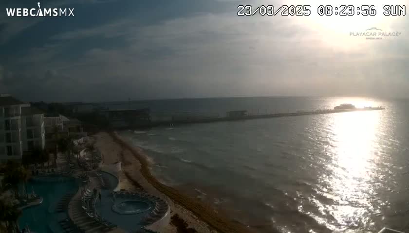 A mostly cloudy sunrise over a calm ocean, showing a beach, resort pool, and long pier.