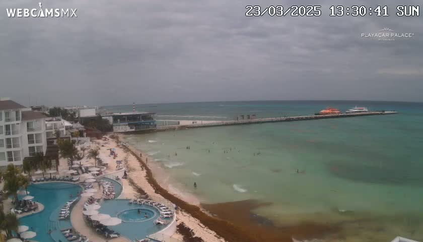 A beach resort with pools overlooks a calm, light-green ocean under an overcast sky, and a pier extends into the water with a ferry docked at its end.