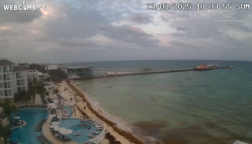An overcast day shows a beach with a resort, swimming pools, and a pier with boats in the distance.