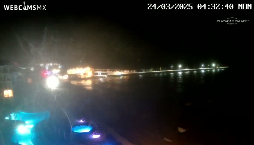 A nighttime, stormy view of a waterfront with illuminated buildings and a body of water, showing  lit pools in the foreground.