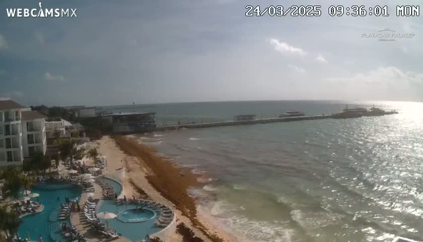 A partly cloudy day reveals a beach with a resort, pools, and a pier extending into the ocean.