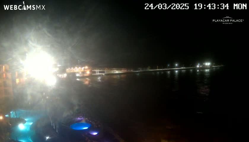 A nighttime view of a brightly lit waterfront area with a body of water in the foreground and a hazy atmosphere, possibly from fog or mist.