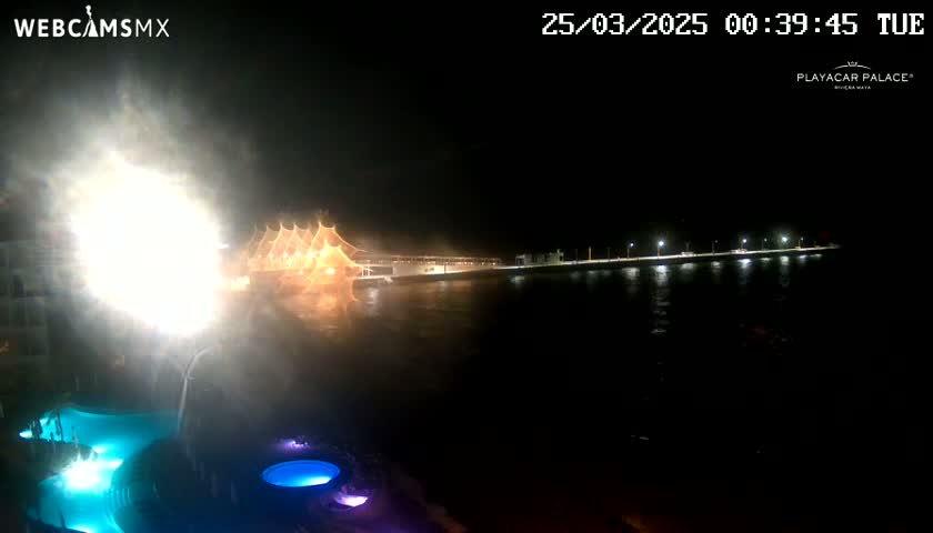 A nighttime view of a waterfront area with brightly lit structures and a large water fountain or firework display, creating a hazy effect over the water.