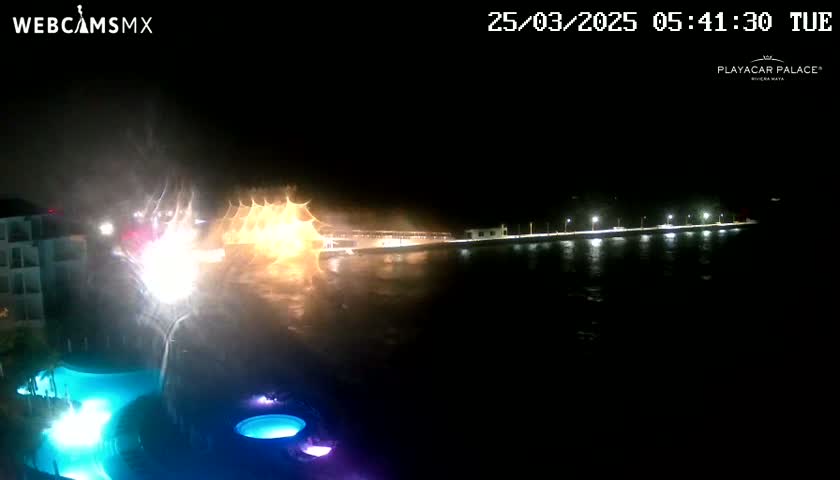 A nighttime view of a resort pool area with illuminated water features, overlooking a rain-swept waterfront with lights lining a distant pier.