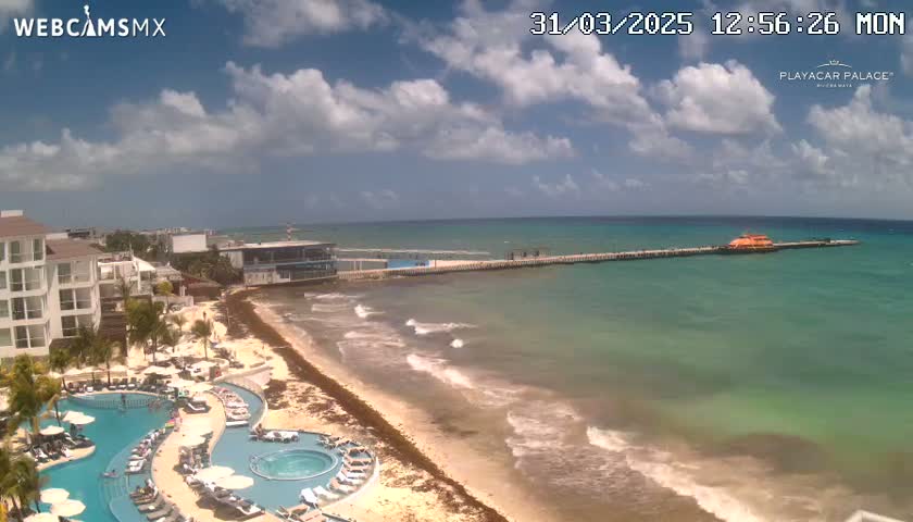 A partly cloudy day shows a beach with a resort, a long pier extending into a calm, turquoise ocean with small waves.