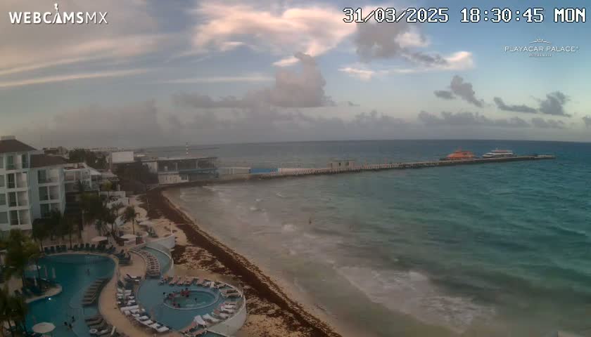 An aerial view of a beach resort with a swimming pool and a pier extending into a partly cloudy ocean with moderate waves.