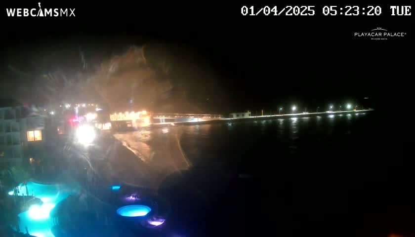 A nighttime view of illuminated pools near a waterfront, with heavy spray or mist obscuring the view of a shoreline and buildings across the water.