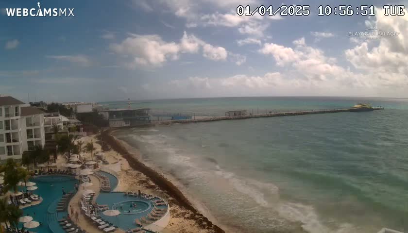 A partly cloudy day shows a beach with a resort, multiple pools, and a pier extending into a turquoise ocean with gentle waves.