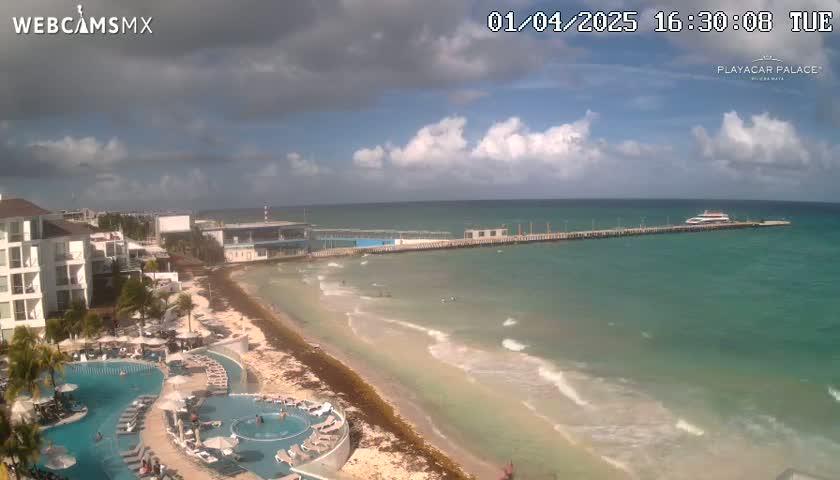 An aerial view shows a partly cloudy day at a beach resort with turquoise water, a long pier with a boat docked at the end, a sandy beach with some seaweed, and a large pool complex next to a building.