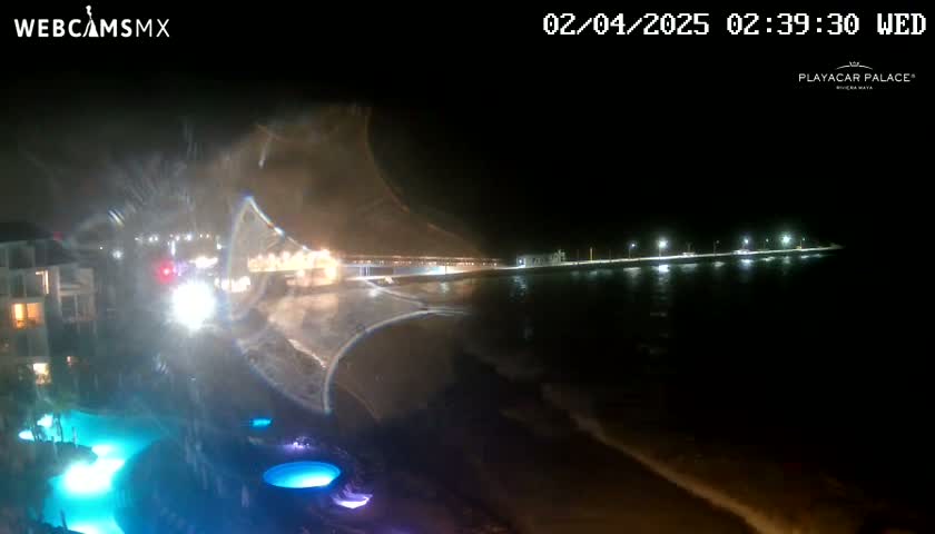 A nighttime coastal scene shows a resort pool lit up in blue, with waves gently lapping the shore near a brightly lit beachfront walkway.