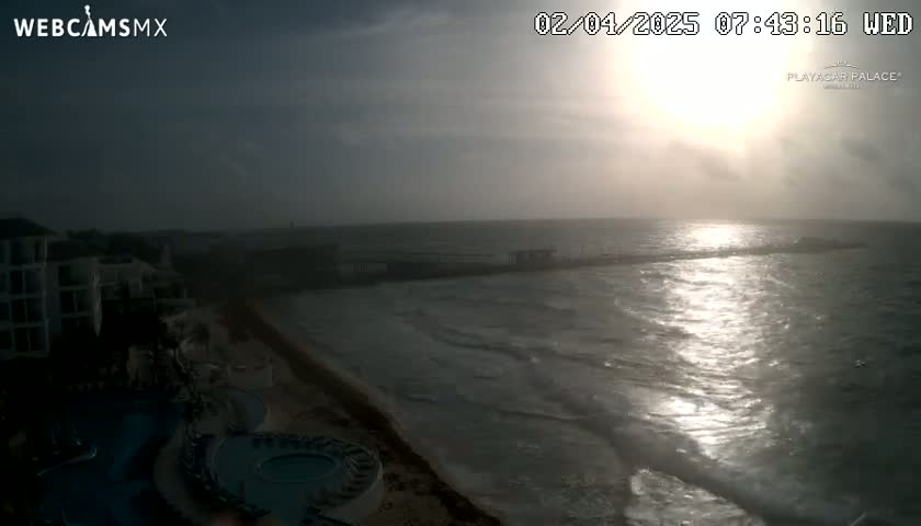 A hazy sunrise illuminates a beach with a resort, a pier, and calm ocean waves.