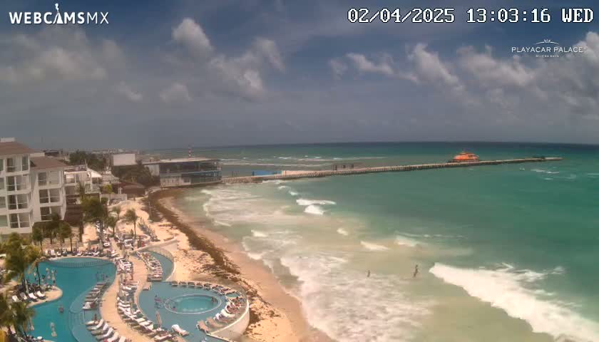 A partly cloudy day shows a resort with a swimming pool complex next to a beach with waves lapping the shore, and a pier extending into a turquoise ocean.