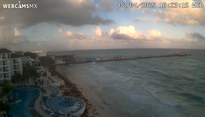 An overcast sky over a resort town with a pier extending into a somewhat choppy ocean, showing a beach and several pools at the resort.