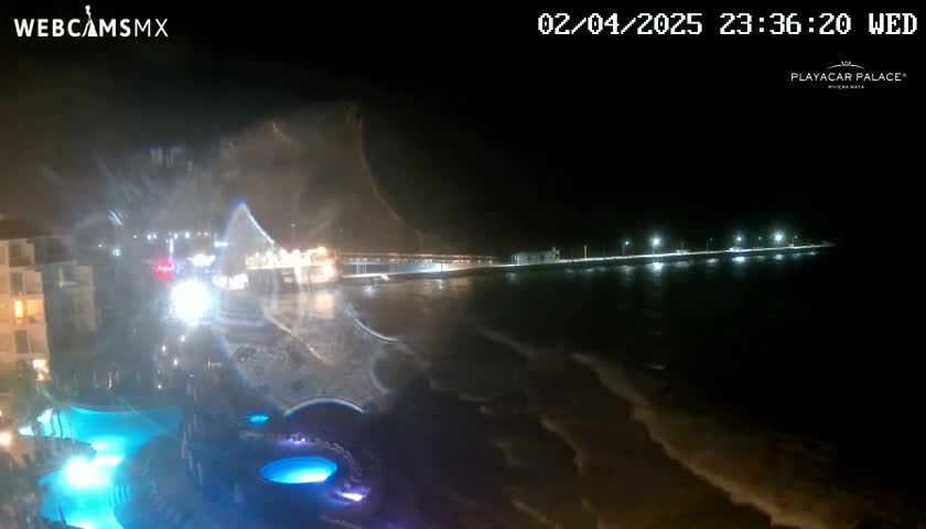 A nighttime view of a coastline shows a building with illuminated pools near a dark ocean with gentle waves under a clear, dark sky.