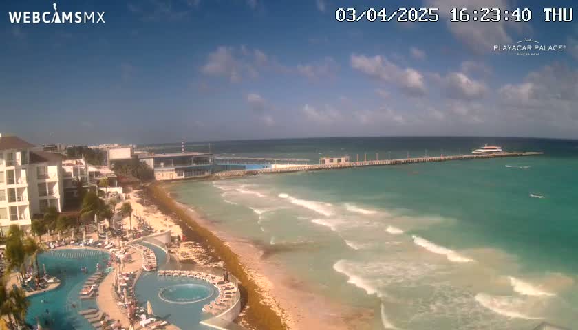 A partly cloudy day shows a beach with turquoise water lapping gently at the shore, a resort with pools, and a pier with a boat moored at the end.