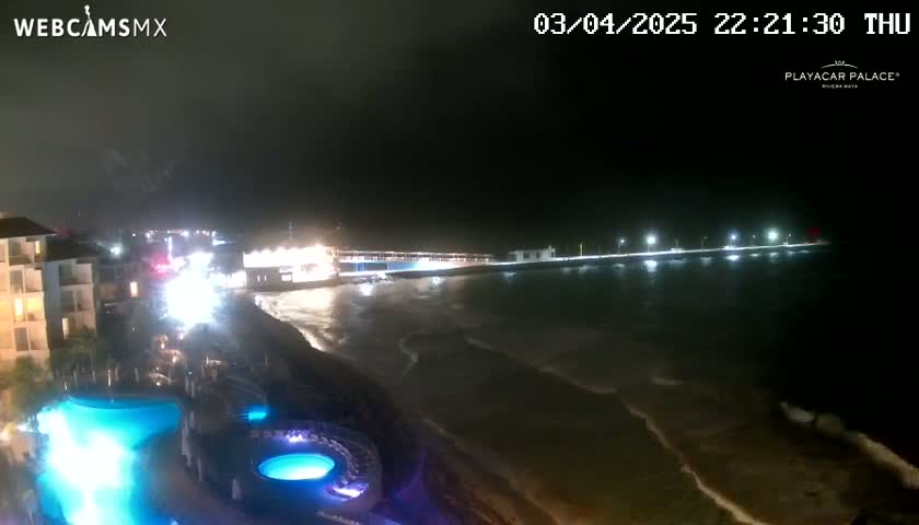 A nighttime view of a resort with illuminated pools overlooking a dark, wavy ocean and a brightly lit pier.