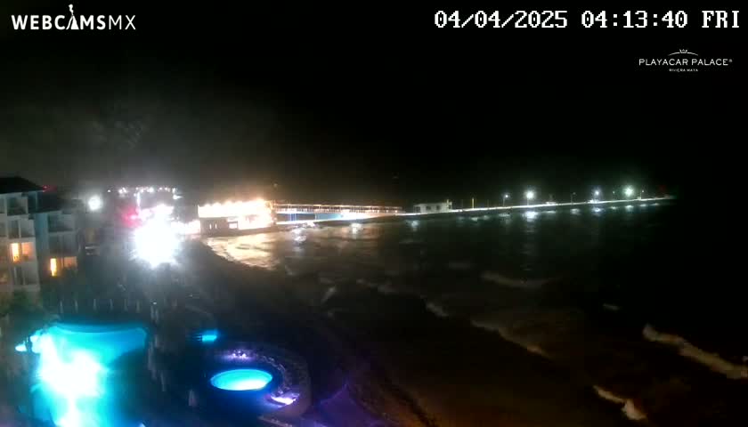 A nighttime view of a coastline shows a resort with illuminated pools and a pier extending into dark, gently waving water.