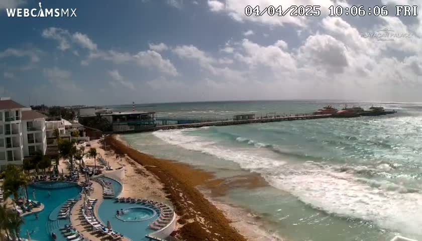 A resort with multiple pools overlooks a beach with seaweed and moderate waves under a partly cloudy sky.