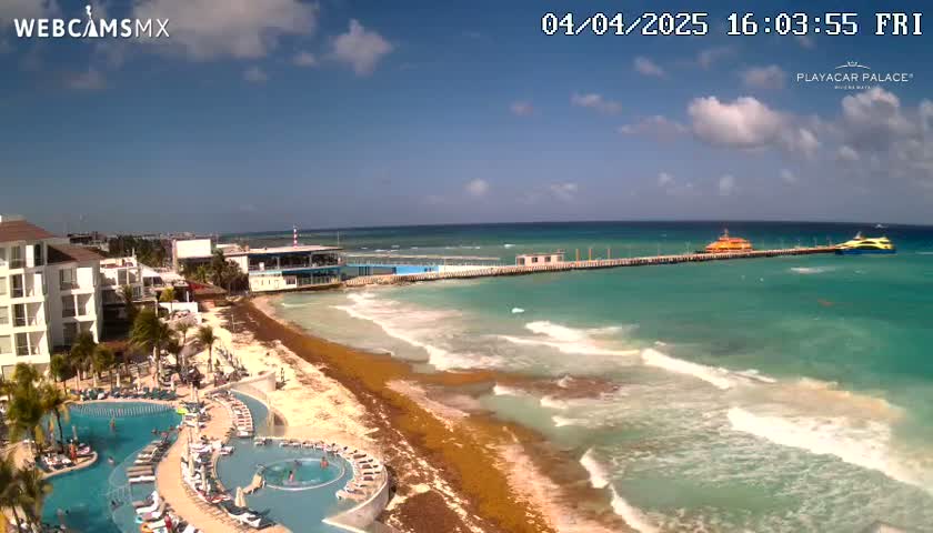 A beach resort with turquoise water and a long pier, featuring two boats and mostly sunny skies with some clouds.