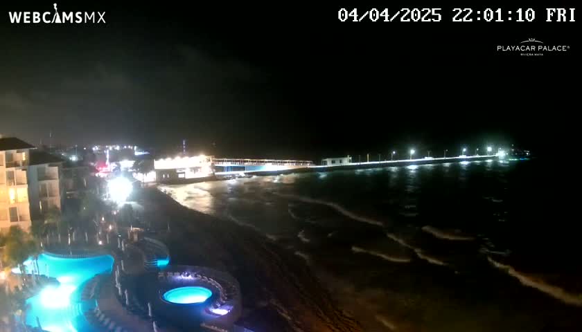 A nighttime view of a resort with illuminated pools, overlooking a dark ocean with small waves under a cloudy sky and a lit pier in the distance.