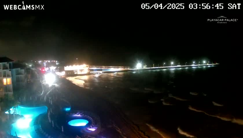 A nighttime view of a beach with a long pier extending into the ocean under partly cloudy skies, featuring illuminated resort pools in the foreground.