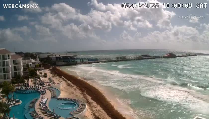 A partly cloudy day shows a resort with pools overlooking a beach with seaweed and a pier extending into a wavy ocean.
