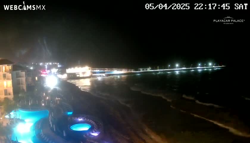 A nighttime view of a resort area shows a beach, ocean with small waves, a long pier with lights, and illuminated swimming pools at the resort.