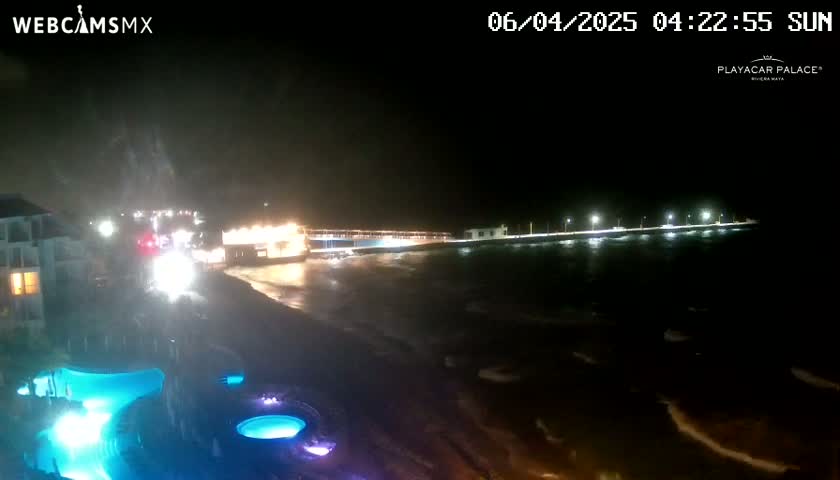 A nighttime view of a beach at the ocean with a pier extending into the water, illuminated by lights, under somewhat stormy conditions.
