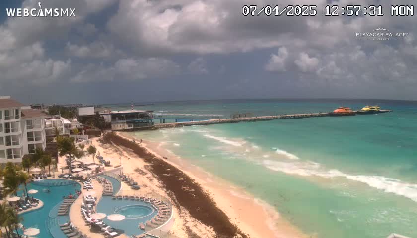 A partly cloudy day shows a resort with multiple pools overlooking a sandy beach and turquoise ocean with a pier and boats in the distance.
