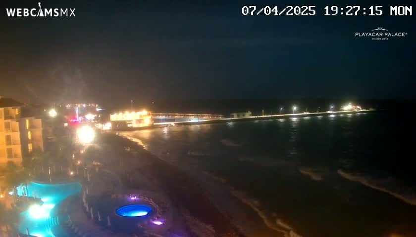 A nighttime view of a beach at a resort, showing a calm ocean with gentle waves lapping the shore, illuminated buildings, and lit pools.