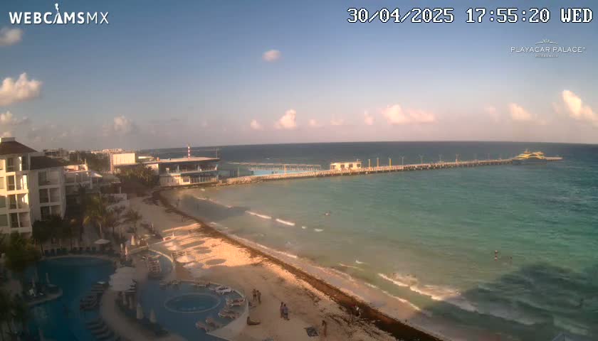A beach with calm turquoise water, a long pier, a resort with a pool, and a partly cloudy sky.