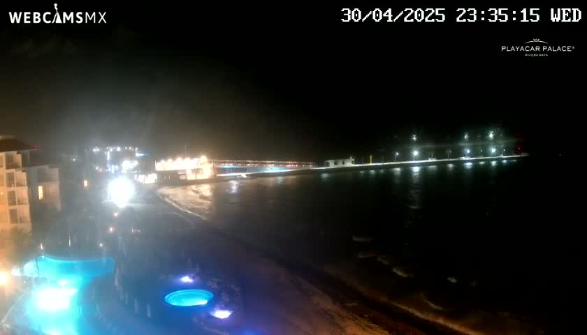 A nighttime view of a beach at a resort, showing a lit pier extending into dark, somewhat choppy water, with a pool and building visible in the foreground.