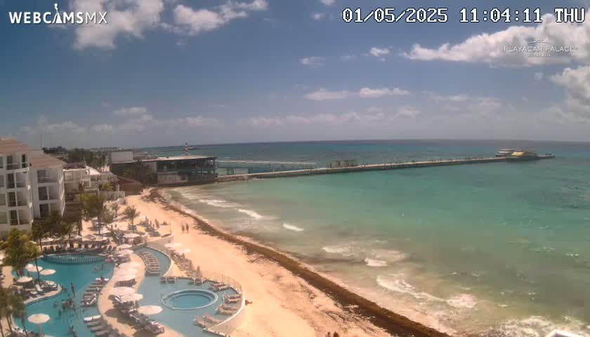 A resort with pools overlooks a beach with a long pier extending into clear, turquoise water under a partly cloudy sky.