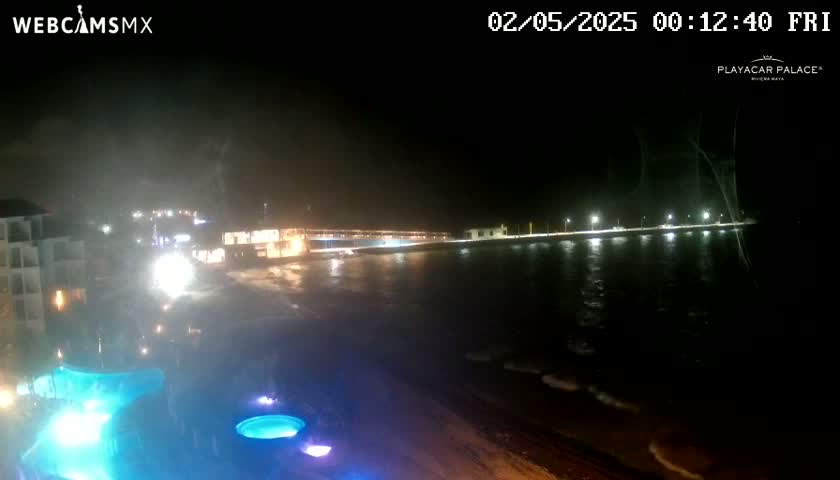 A nighttime view of a beachfront resort with a lit-up boardwalk and ocean waves under stormy conditions.