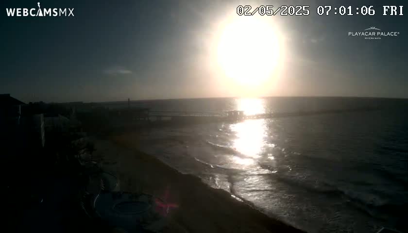 A bright sunrise over a calm ocean reflecting the sun's light, with a sandy beach and shoreline visible in the foreground.