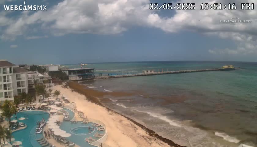 A partly cloudy day shows a resort with pools overlooking a beach with seaweed, a pier extending into a calm turquoise ocean.