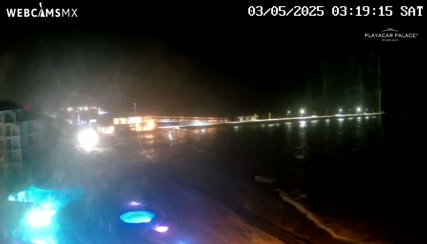 A nighttime view of a shoreline with buildings and lights, showing a partly cloudy and windy night with a somewhat choppy body of water.