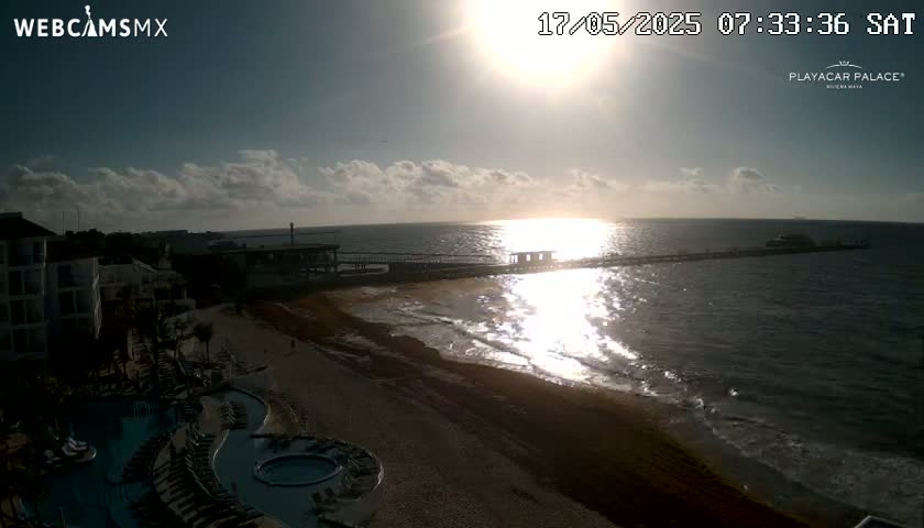 A partly cloudy sunrise over a beach with a long pier extending into the ocean, showing a resort with pools in the foreground.
