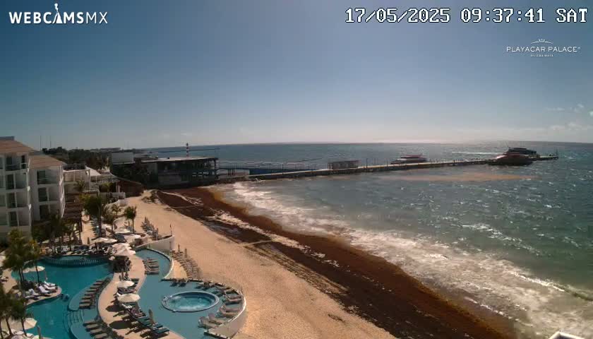 A resort with multiple pools overlooks a beach covered in seaweed, with boats docked at a pier in the clear, sunny ocean.