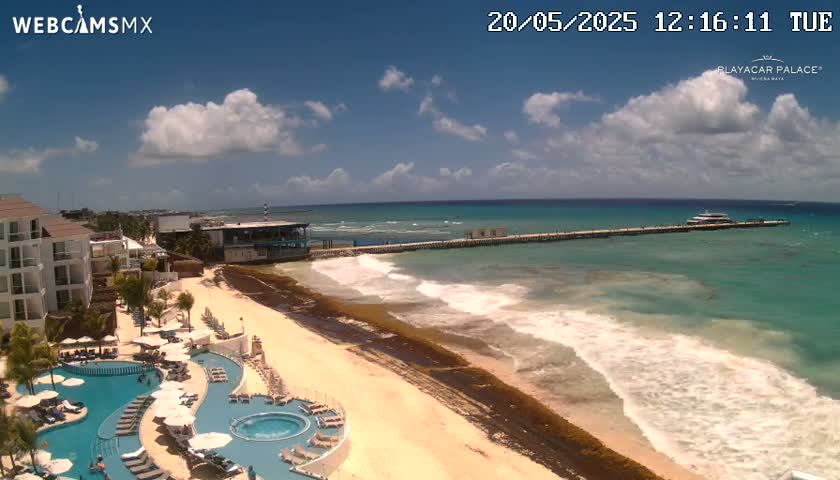 A resort with multiple pools overlooks a sandy beach with seaweed along the shoreline and a pier extending into a turquoise ocean under a partly cloudy sky.