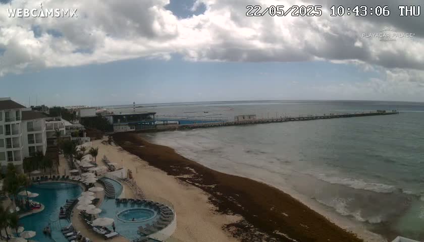 A partly cloudy day shows a beach with seaweed, a resort with pools, and a long pier extending into a calm ocean.