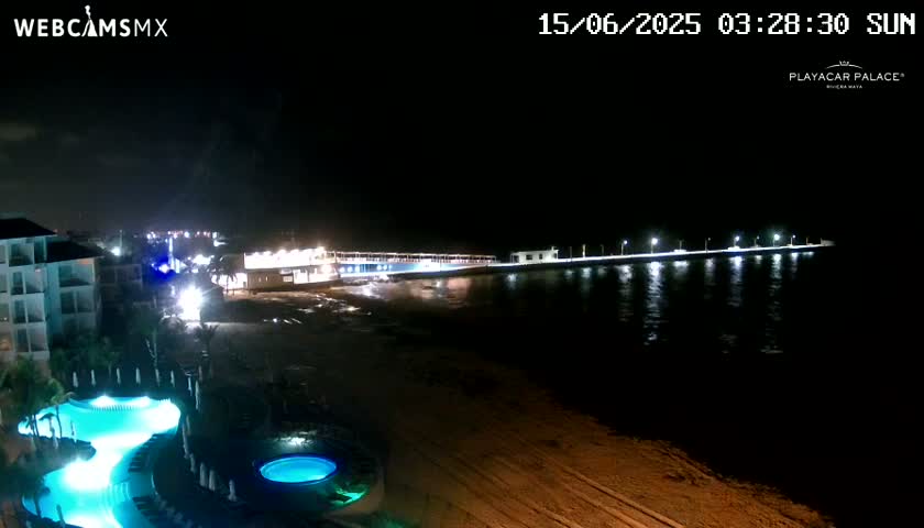 A nighttime beach scene shows a brightly lit pier extending into dark water, with a resort pool and buildings visible in the foreground under a clear sky.