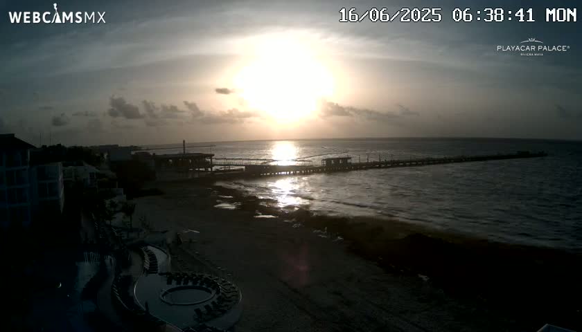 A beach scene at sunrise shows a long pier extending into a calm ocean, with buildings and a sandy beach visible in the foreground.