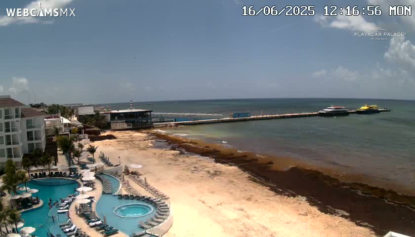 A resort with multiple pools overlooks a sandy beach with seaweed along the shoreline, where two boats are docked at a pier under a partly cloudy sky.