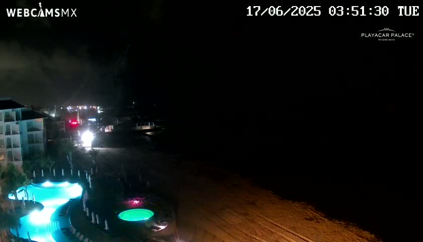 A nighttime view of a resort area with illuminated pools and a dark beach under cloudy skies.