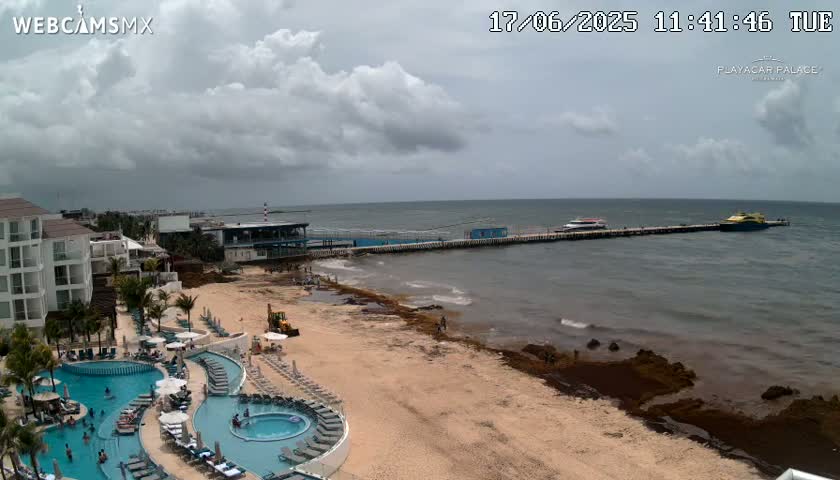 A resort with swimming pools overlooks a beach with seaweed and a pier where boats are docked, under a partly cloudy sky.